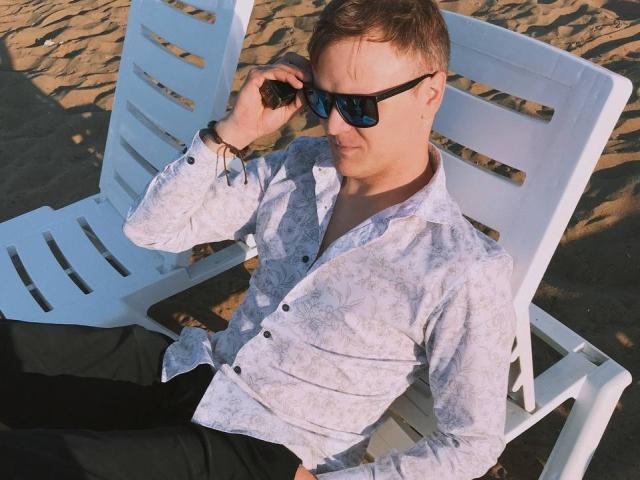 ChillOutt é um rapaz com cabelo castanho curto, usando óculos de sol pretos, uma camisa florida e calças pretas, sentado em uma cadeira de praia branca..