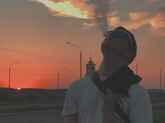 Rilassati con gli occhiali da sole, la camicia bianca, la sciarpa nera e il cinturino, con la testa inclinata all'indietro, mentre ammiri il tramonto arancione. Il cielo è pieno di nuvole arancioni e gialle, e sullo sfondo si vedono i lampioni..