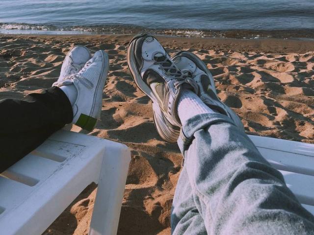 Las piernas de ChillOutt, con zapatillas blancas y grises, pantalones negros y calcetines grises, descansan sobre una silla de plástico blanca, con vistas a una playa de arena y agua azul..