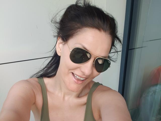 JoliDanielle mit dunklem Haar in einem unordentlichen Pferdeschwanz, trägt ein grünes Tanktop und goldene Aviator-Sonnenbrillen, lächelt mit sichtbaren Zähnen und zeigt Oberkörper und Gesicht..