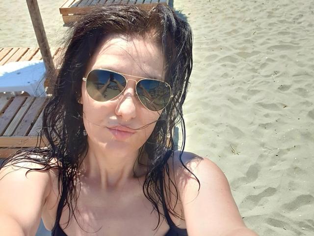 JoliDanielle with wet black hair, wearing gold aviator sunglasses and a black top, showing her small tits and pouty lips, on a sunny sandy beach.