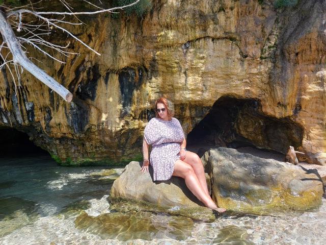 CurvySweetGirl met een groot achterwerk en een lichtroze bloemenjurk, zittend op een rots aan het water, met een zonnebril op, benen gekruist, bruin haar, een rotsachtige klif op de achtergrond, een zonnige dag, de waterlijn..