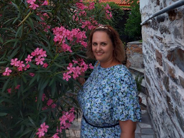 CurvySweetGirl met een roze bloemenstruik, gekleed in een blauwe bloemenjurk, een zwarte riem, lachend, met bruin haar, een hoofdband, staand naast een stenen muur. Borsten licht zichtbaar onder de jurk. Buitenomgeving..