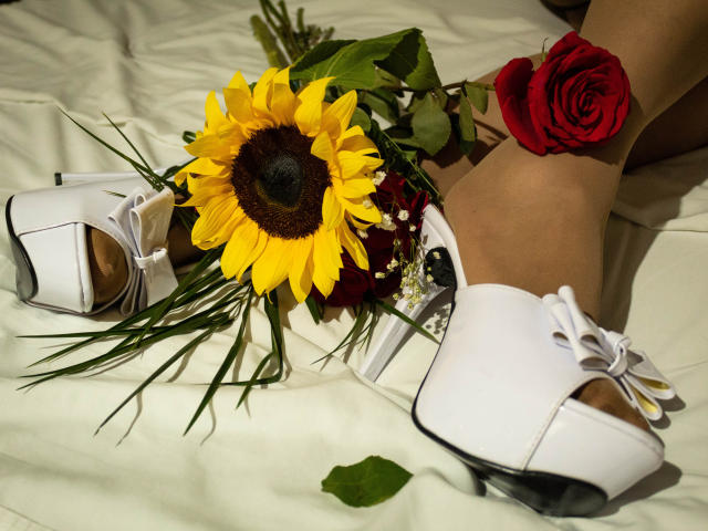 DianeOasis met blond haar, gekleed in zwart kant, die haar borsten bedekt met één hand, een gouden ketting, rode lippenstift, en die een boeket houdt met zonnebloemen en rode rozen, in een zwak verlichte kamer..