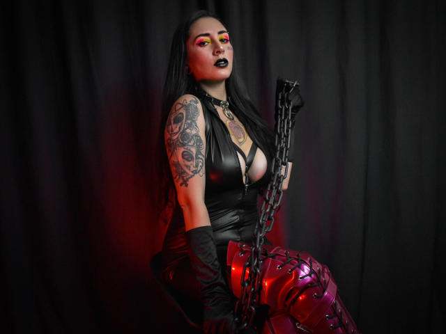 EmillyManson, vêtue de cuir noir, avec des reflets rouges, des gants noirs, une chaîne, un bras tatoué et un maquillage gothique, tenant une chaîne, sur un fond sombre, avec un éclairage rouge, montrant ses seins et ses fesses..