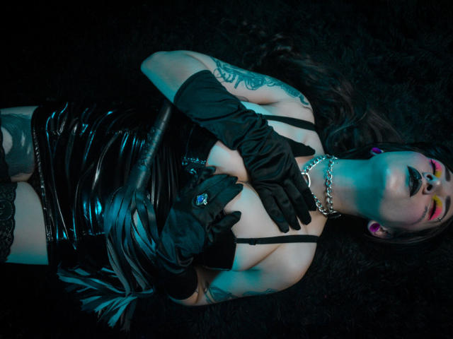 EmillyManson lying on dark fur, wearing black latex dress, gloves, and chain necklace, with tattoos and colorful face paint, covering her tits with black-gloved hands.