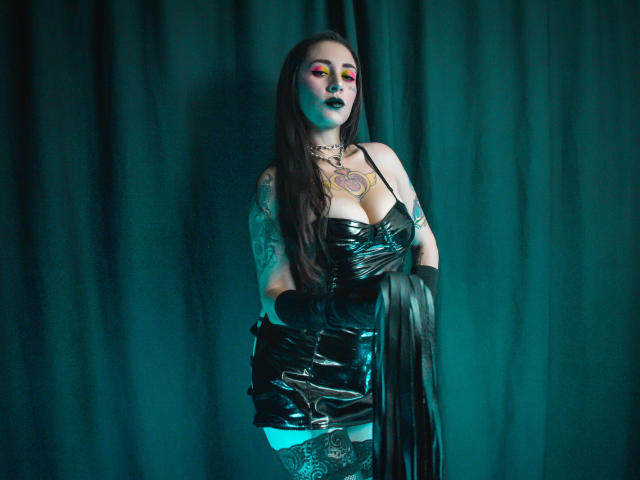 EmillyManson, vêtue d'une robe en latex noir, tenant un fouet en cuir, avec un bras tatoué, du fard à paupières rose, un éclairage bleu-vert, des cheveux foncés, de grandes seins, portant des gants, avec un fond de rideau vert..