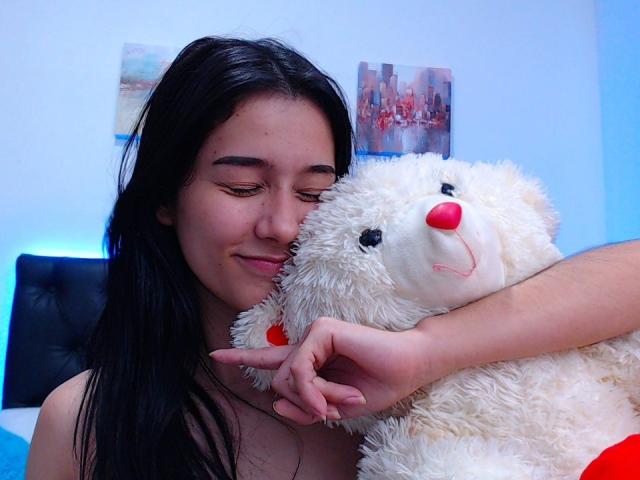 DeisyWest with long black hair, cute smile, and closed eyes, hugging a fluffy white teddy bear with a red nose, in a bedroom with blue lighting.