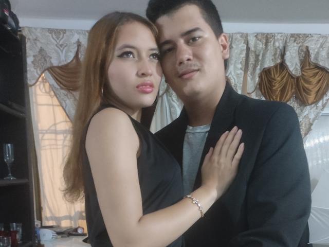 JakeAndMegan, con i capelli castani e le labbra piene, indossa un abito nero e ha una mano sul petto di JakeAndMegan, che indossa una giacca nera, mentre si trovano in una stanza con tende elaborate..