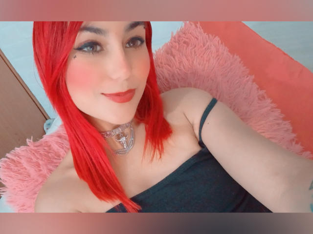 AmyStreetwise69 with red hair, wearing a black strap top, pink fluffy pillow, and a silver necklace, smiling at the camera with red lipstick and a small mole above her lip.