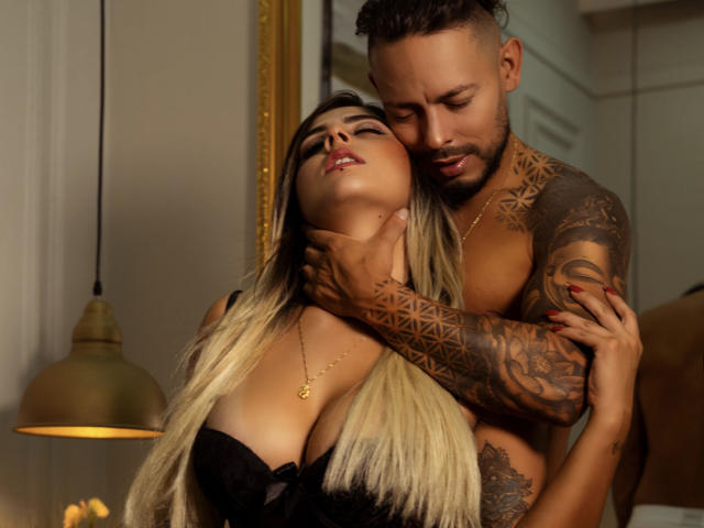 KendraWaltonJeffLuthor, com cabelo loiro, seios grandes em lingerie preta, sendo abraçada por KendraWaltonJeffLuthor tatuada, com cabelo curto, olhando para baixo, em uma pose quente, íntima e sensual..