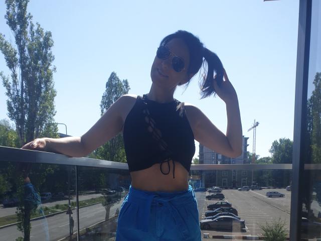 JoliDanielle with sunglasses, black crop top showing her small tits, blue pants, and hand in ponytail, leaning on glass railing, sunny street background.