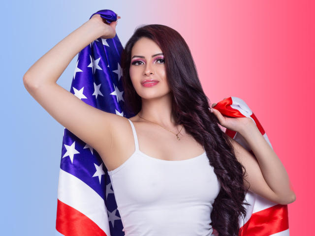 HannaRobertss with long brown hair, white tank top, holding American flag, blue left, pink right background. She's smiling, flag over shoulder, right arm raised. Boobs visible, cute.