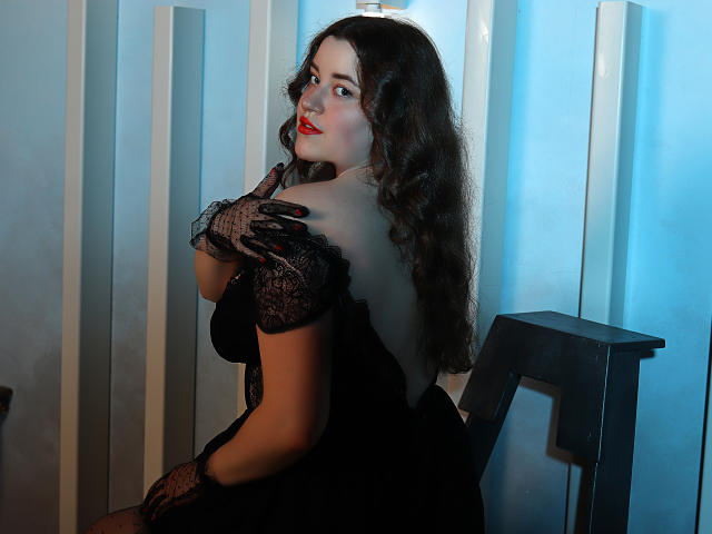 PolinaGrace mit welligem, dunklem Haar, rotem Lippenstift, schwarzen Spitzenhandschuhen und einem schwarzen Kleid, sitzt auf einem Stuhl, blickt über ihre Schulter und zeigt ihr Gesäß..