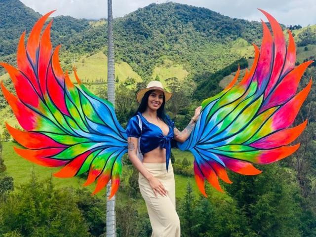 Danna Stevens, avec des ailes arc-en-ciel colorées, un haut bleu à nouer, un pantalon beige, un chapeau de paille, souriante, tenant une aile, avec un arrière-plan de montagnes vertes, et un tatouage sur le bras droit..