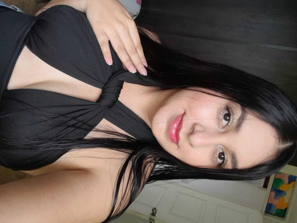 Luiisa69, com cabelo preto, lábios rosa e olhos castanhos, vestindo uma blusa preta com um nó, mostrando os seios, segurando a blusa, sorrindo, em um selfie..