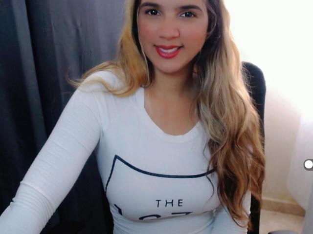 Wendy, con cabello rubio largo, camisa blanca de manga larga y lápiz labial rojo, sonriendo a la cámara, mostrando sus pechos de tamaño medio, sentada en una silla negra..