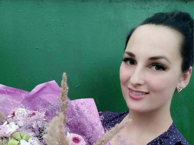 BabeBeautiful com cabelo escuro preso em um rabo de cavalo, segurando um buquê roxo, sorrindo para a câmera, vestindo um vestido escuro com bolinhas, fundo verde, pequenos brincos de ouro..