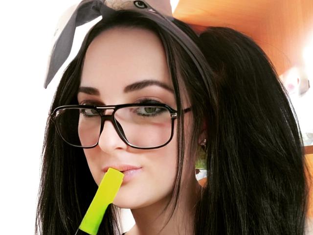 Een mooie vrouw met zwarte vlechtjes, een bril en een konijnenoorhoofdband, die aan een felgroene rietje zuigt, waarbij haar volle lippen en subtiele make-up zichtbaar zijn. Haar borsten zijn niet zichtbaar..
