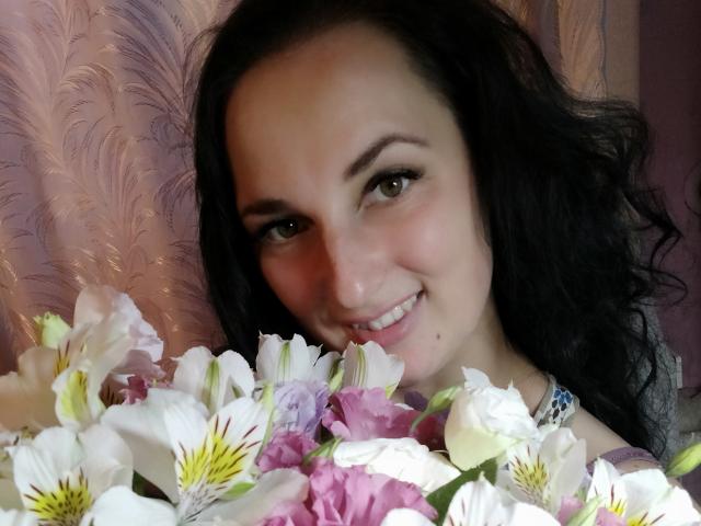 Uma mulher linda, com cabelos cacheados escuros, sorrindo, com o rosto parcialmente coberto por flores brancas, rosa e roxas, olhos verdes, pele clara, maquiagem discreta, usando um colar azul..