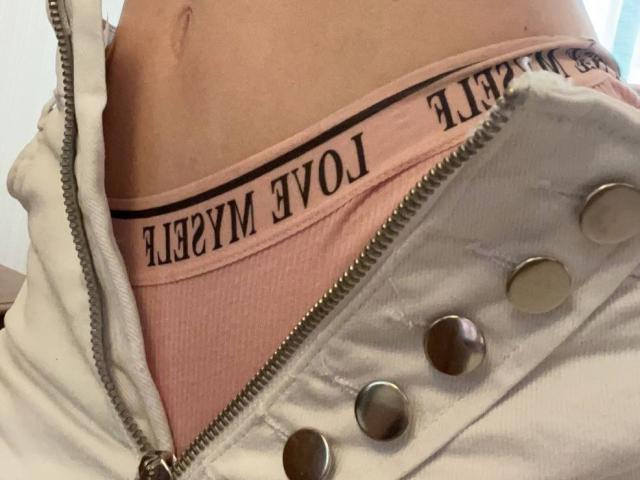SelinaRose's pink underwear is visible under a white zip-up jacket with metal buttons, showing her lower belly and waist. Her belly button is slightly visible.