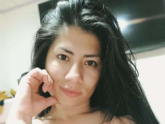 GenesisThonson tiene el cabello largo y negro, ojos marrones y labios rosados ligeramente separados, con el mentón apoyado en la mano. Está mirando directamente a la cámara con una sonrisa sutil..