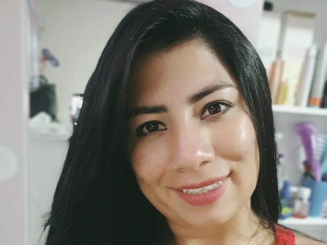 GenesisThonson, avec ses cheveux noirs, ses yeux marrons et un sourire, montrant ses lèvres pulpeuses et un léger espace entre ses dents, porte un haut rouge, sur une photo d'elle où l'on aperçoit un léger éclat sur son visage..