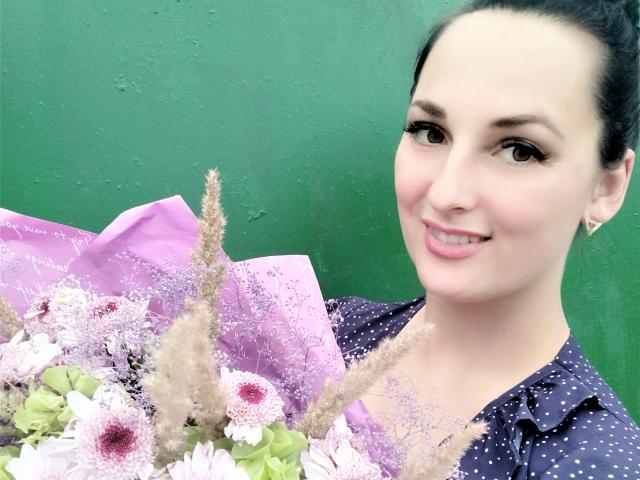 Eine wunderschöne Frau mit dunklem Haar zu einem Dutt gebunden, lächelt in die Kamera, hält einen Strauß aus rosa und weißen Blumen mit grünen Stielen und trägt ein lila Hemd mit Punkten..
