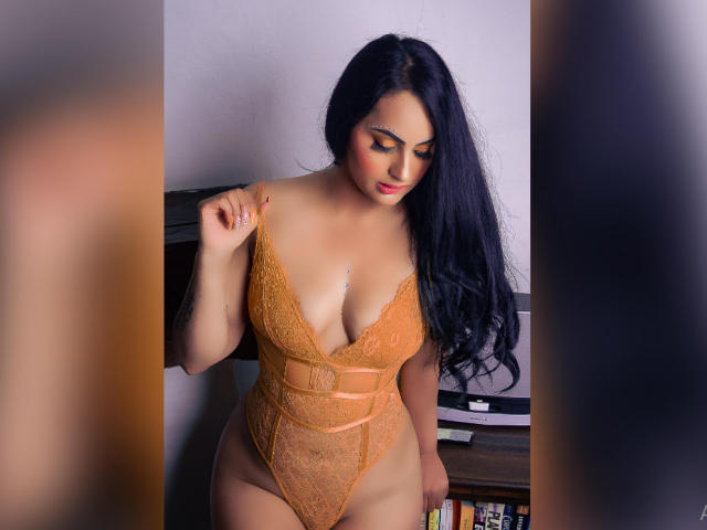 MartinaMendezz, con cabello negro largo, vestida con un body de encaje naranja, mostrando sus grandes senos y su trasero curvilíneo, mirando hacia abajo con una expresión suave, con una mano en el hombro..