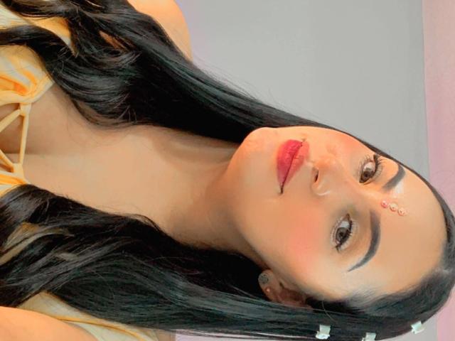 MartinaMendezz tem cabelos pretos longos, batom vermelho e um pequeno bindi na testa. Ela está vestindo uma blusa amarela, com os seios levemente visíveis. As nádegas dela não estão à mostra..