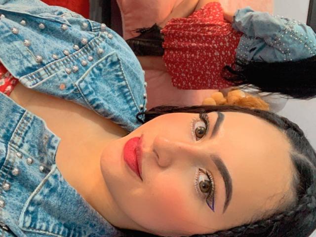 MartinaMendezz tiene una cara linda con ojos marrones, lápiz labial rosa y cabello trenzado. Lleva una chaqueta vaquera azul con tachuelas plateadas, mostrando ligeramente sus senos..