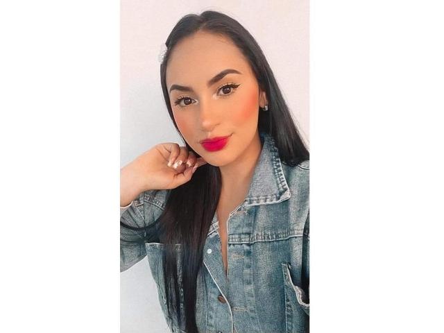MartinaMendezz mit langen, schwarzen Haaren, die eine Jeansjacke trägt, roten Lippenstift und auffälligem Eyeliner, ihre Hand in der Nähe ihres Ohrs, blickt mit einem leichten Lächeln in die Kamera..