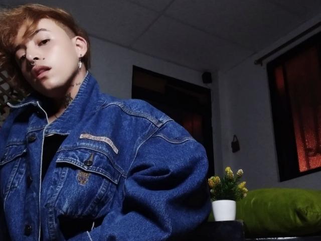 YeiSela met kort bruin haar, gekleed in een blauwe denimjas, kijkt opzij, met oorringen, een groene kussen, een kleine potplant, gele bloemen en een donkere kamer..