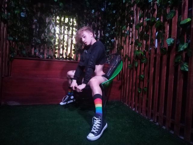 YeiSela, met blond haar, een zwart topje, regenboogkousen en zwarte sneakers, zit in een squat op een groene stoel en toont haar kale, glanzende achterkant, in een houten, begroeide, zwak verlichte omgeving..