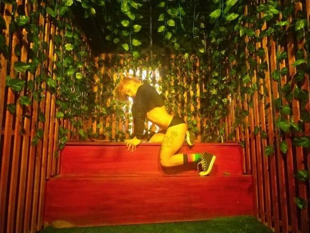 YeiSela is on red stairs, kneeling with ass up, wearing a black crop top, green socks, and green-and-black sneakers, with leafy vines hanging around.