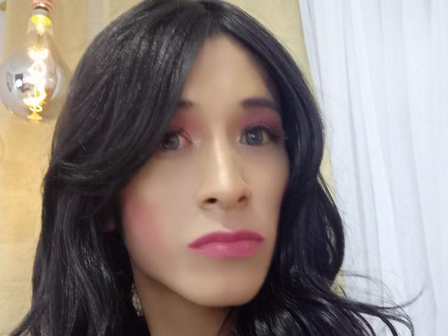 ThomasParker69 a les cheveux noirs et ondulés, du fard à paupières rose et du rouge à lèvres rose. Son visage est au centre de l'image, avec une ampoule visible en arrière-plan. Ses seins ne sont pas visibles..