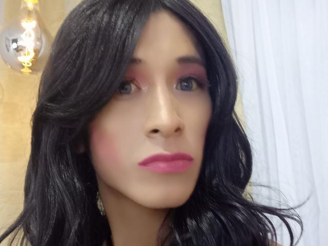ThomasParker69 with black wavy hair, pink eyeshadow, and pink lipstick, has a light skin tone and blue eyes, showing a neutral facial expression. Light bulb in background.