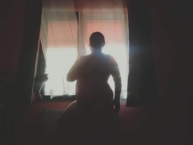 CurlyCherry, silhouetteée, se tient nue, dos à la caméra, près d'une fenêtre lumineuse avec des rideaux transparents, une main sur la hanche, une lumière douce soulignant ses courbes et son dos, dans une pièce sombre..
