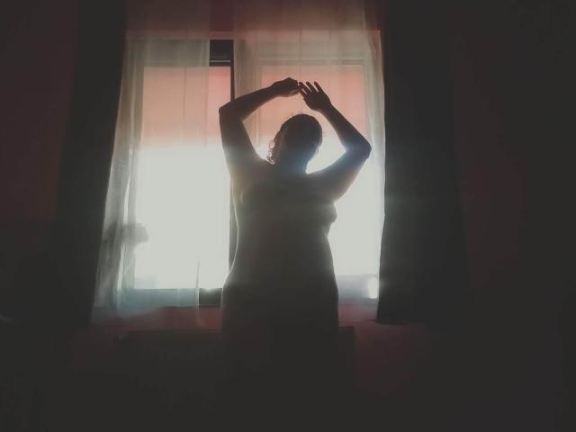 CurlyCherry, con i capelli ricci e la silhouette delineata, con le braccia alzate, in piedi di fronte a una finestra luminosa, illuminata di luce posteriore, creando una figura ombrosa con il seno visibile e i fianchi prosperosi, in una stanza poco illuminata..
