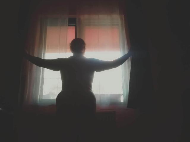 Silhouetted CurlyCherry staat met armen gespreid, met de rug naar het raam, waarbij het licht dat door de gordijnen schijnt, haar gespierde rug en billen accentueert. Het zachte licht creëert een subtiele, sexy gloed..