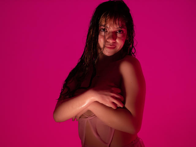ElizabethBlizz mit nassen, braunen Haaren, die ihre Brüste gegen einen rosa Hintergrund verdecken, blickt mit einem leichten Lächeln in die Kamera. Sie trägt ein durchsichtiges, rosa Oberteil..