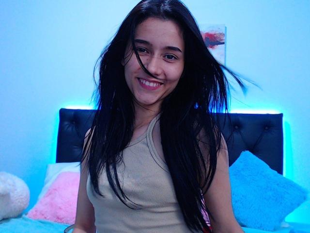 DeisyWest with long black hair, smiling, wearing a beige tank top, showing small tits, sitting on a bed with blue and pink pillows, and a black tufted headboard.