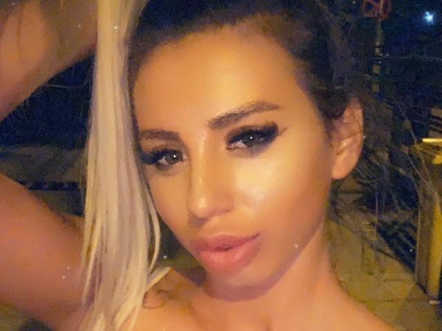 LisaaKiraa mit blonden Haaren, starkem schwarzem Eyeliner und glänzenden rosafarbenen Lippen, ihr Gesicht strahlend im warmen Licht, ihre Hand erhoben zum Kopf. Ihr Hintern ist nicht sichtbar..