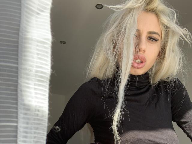 LisaaKiraa with long, messy blonde hair, black turtleneck, and pouty pink lips, standing near a white, ribbed curtain. Her makeup is bold with thick eyeliner.