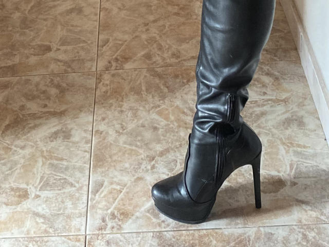 ArleenHunter's black leather high-heeled boot with a zipper, standing on beige marble tile. Boot is shiny, stiletto heel is tall, and her leg is slightly bent.