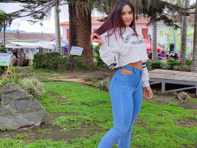 JessicaFit, con lunghi capelli rosso scuro, indossa una felpa bianca a maniche corte e jeans blu a vita alta, in piedi in un parco erboso, mostrando la sua vita sottile e i suoi fianchi formosi..
