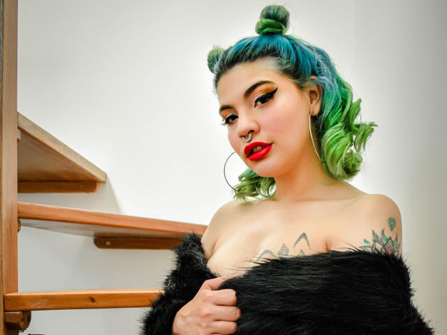 AndiChin con i capelli verdi raccolti in chignon, rossetto rosso, pelliccia nera, orecchini a cerchio, piercing al naso, tatuaggi e eyeliner spesso, che tiene in mano della pelliccia nera e mostra il seno..