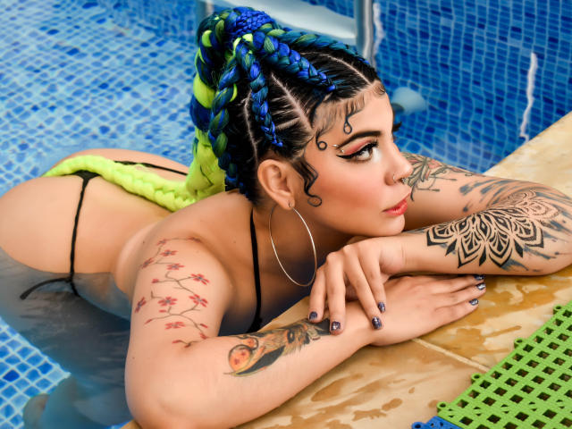 AndiChin, avec ses cheveux tressés bleu-vert, son string noir, son bas de maillot de bain en crochet jaune néon, allongée dans la piscine, montrant ses tatouages, de grandes boucles d'oreilles, regardant sur le côté, fesses en l'air, bras sur le bord jaune de la piscine..