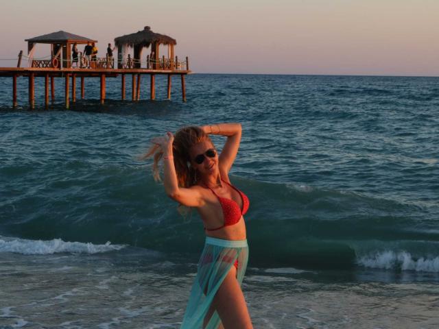 LadyLea with red bikini top, showing her boobs, and sheer blue skirt, standing in the ocean, hands in her blonde hair, wearing sunglasses, at sunset.