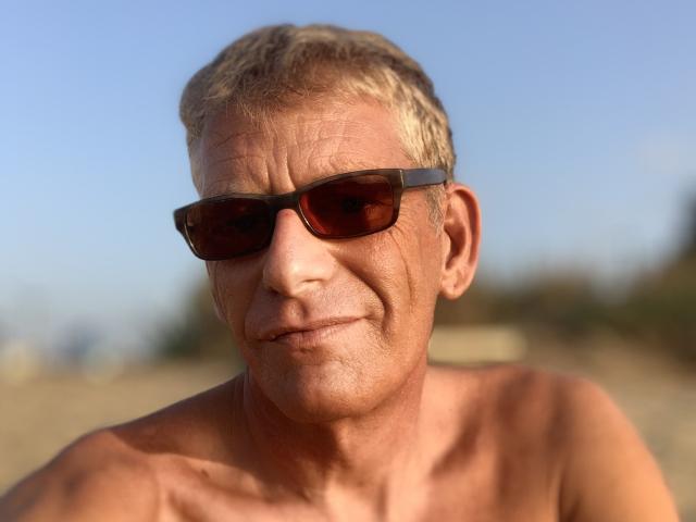 SergioEsp, zonder shirt, met gebruinde huid, draagt donkere zonnebril, heeft kort grijs-blond haar en een lichte glimlach, en kijkt recht in de camera..